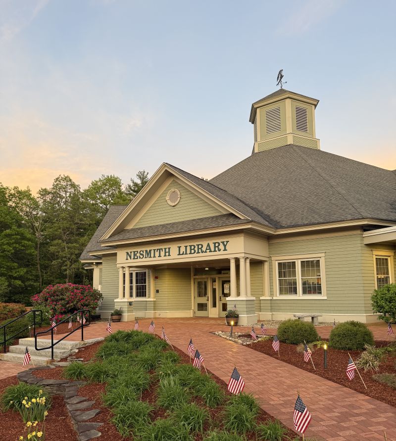 Exterior of Nesmith Library at dusk, a light green building with white columns and a cupola, surrounded by small American flags lining the front walkway