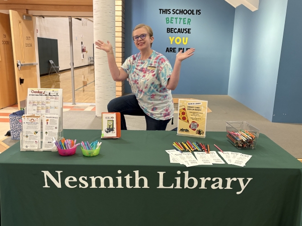Azra, Head of Youth Services, smiles behind a Nesmith Library table with printed handouts and a coloring page for kids.
