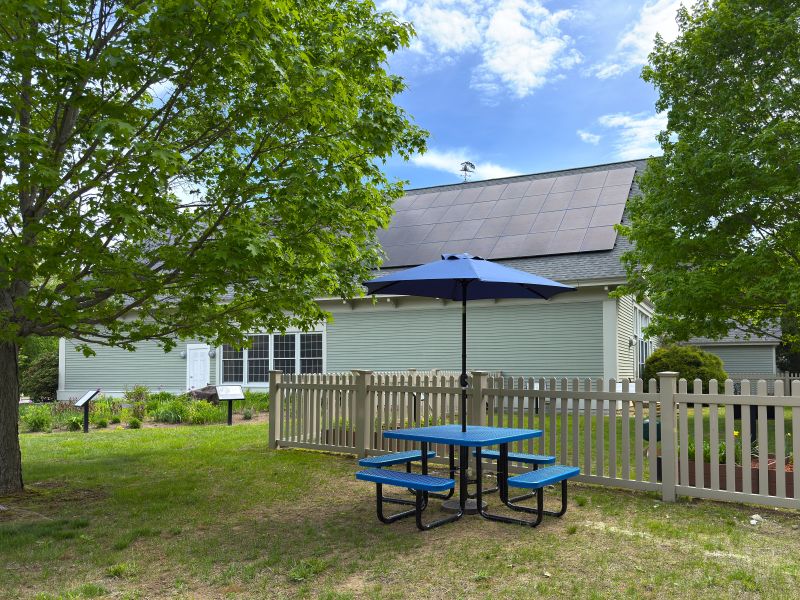 A blue picnic table with a blue umbrella sits outside the Windham Public Library, next to a white picket fence surrounding the Story Garden.